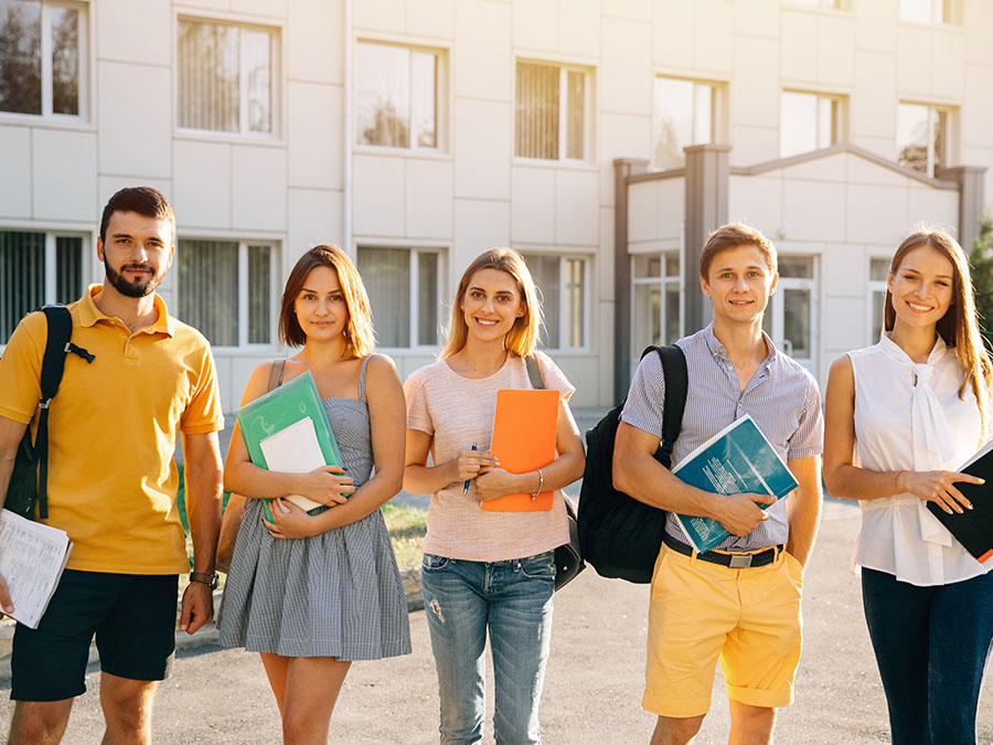 Eine Gruppe junger Erwachsener stehen vor einem Schulgebäude. Die Sonne scheint.