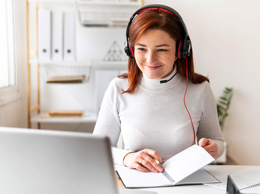 Eine rothaarige Frau sitzt lächeln vor ihrem Laptop und trägt ein Headset.